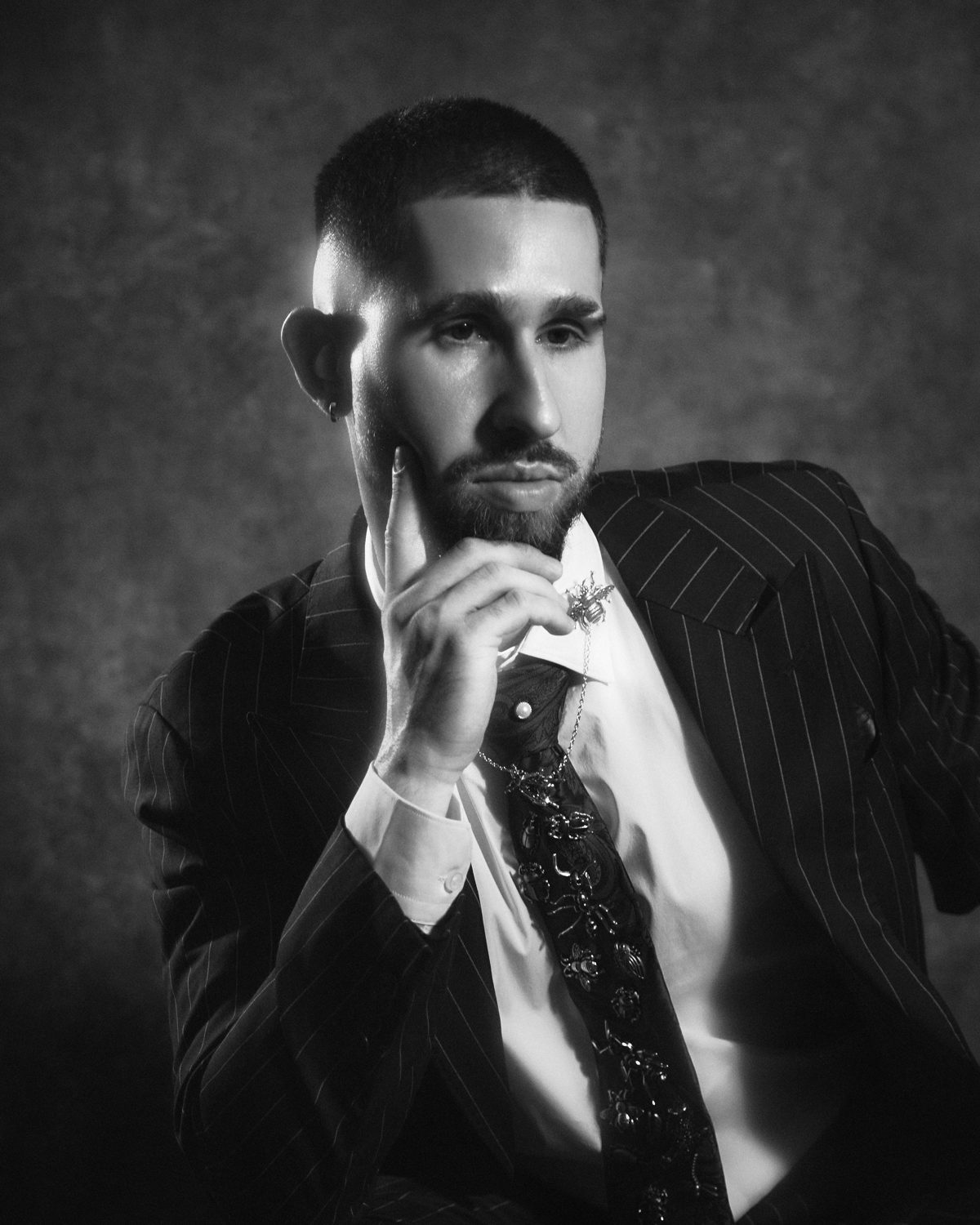 Black and white portrait of Edoardo Cantiello in a pinstripe suit and insect-embroidered tie, a finger resting on his cheek