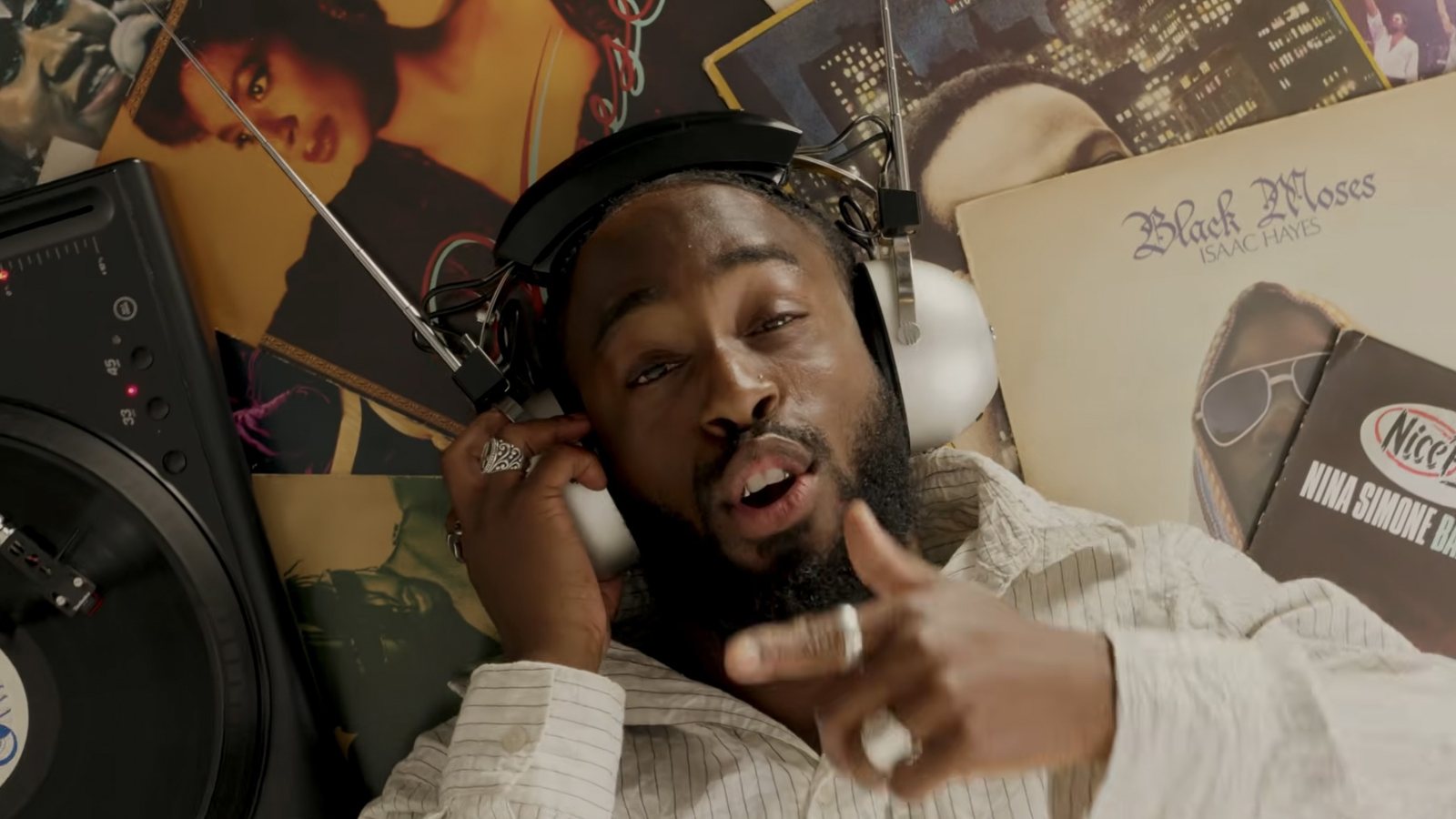 Kadeem Tyrell lies on a floor of vinyl records wearing big headphones, pointing at the camera