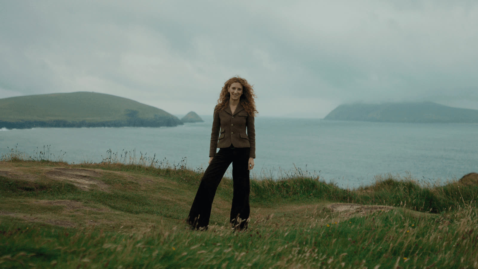 A woman with red curly hair stands on a grassy clifftop above the sea in County Kerry